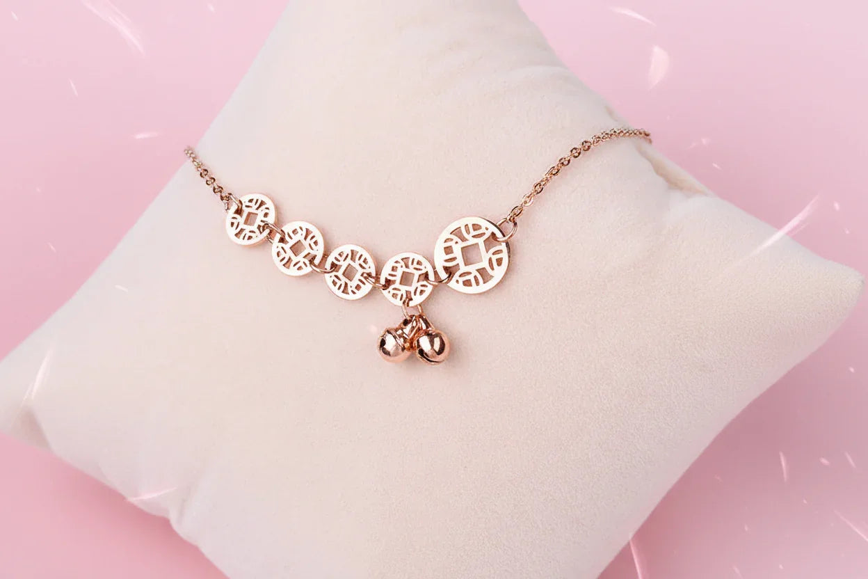 Rose gold bracelet with circular and bell-shaped charms on a light pink surface.