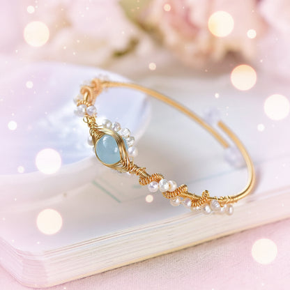 Gold bracelet with pearls and a blue gemstone on a white surface with flowers in the background