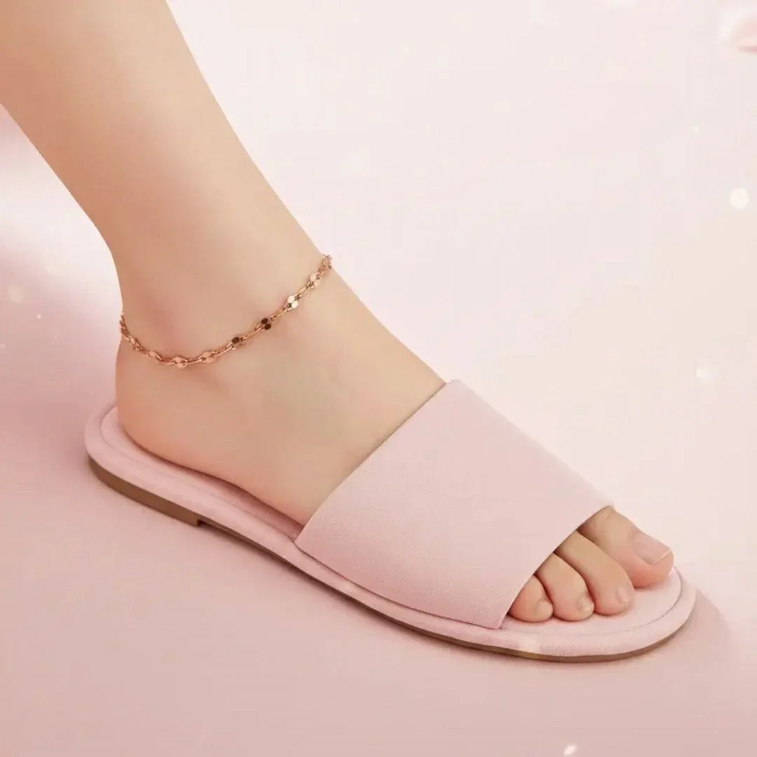 Pink sandal worn on a foot with a gold anklet against a light pink background